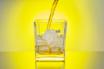 Whiskey is poured into a transparent glass with ice against the background of a yellow light source