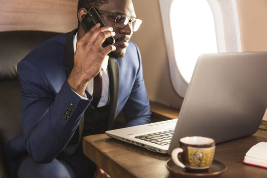 Young attractive and successful African American businessman with glassies talking on the phone and working while sitting in the chair of his private business plane