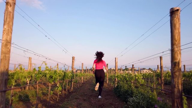 Bella ragazza con maglia rosa e pantaloni neri corre felice in mezzo ai vigneti in campagna.