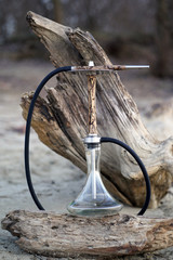 Beige wooden hookah with lightning patterns from an electric current with a clean transparent bowl with a hose with metal elements on a log in the forest on the background of a fallen tree.