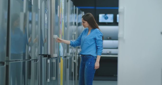 Young sexy brunette woman in shirt to open the door of the refrigerator in the appliances store and compare with other models to buy the new house