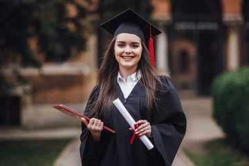 Female student graduate is standing in university hall in mantle, smiling and looking at the camera...