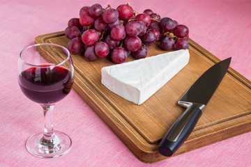 Glass of red wine and a triangular piece of Brie cheese, red sweet grapes, chef knife on a brown wooden cutting board. Soft cheese covered with edible white mold.