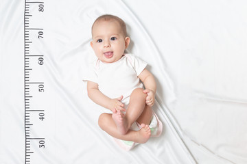 A five month baby in white clothes lying on a bed on which a measuring ruler for growth is drawn. shows the tongue from her mouth and teased