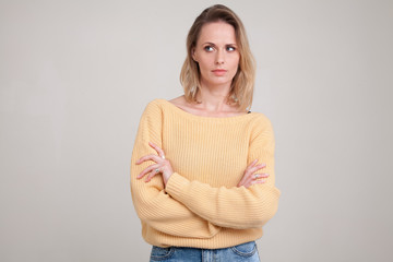Fototapeta premium waist-up portraite of blonde woman with puzzled face expression looks aside with her arms folded. wearing yellow sweater. poses against grey background. face expression, body language