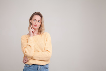 portrait of blonde woman with puzzled face expression,keeps her finger on cheek, looks aside. wearing yellow sweater.poses against grey background.free space for your promotional text or advertisement