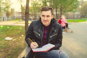 Obraz premium Man with a notebook in his hands in the Park. Male, student is writing her ideas and thoughts into the notebook sitting on the bench in the park 