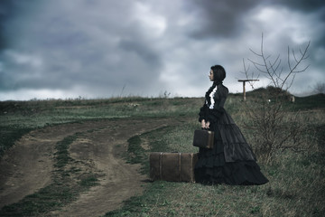Outdoors portrait of a victorian lady in black sitting alone on the road with her luggage. - obrazy, fototapety, plakaty