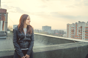 Fototapeta na wymiar Woman in trendy jacket on roof edge. Portrait of young female in leather jacket, standing on the roof of at sunset