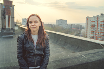 Woman in trendy jacket on roof edge. Portrait of young female in leather jacket, standing on the roof of at sunset