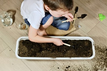 A child with garden tools doing work in pots of earth, sitting next to the side of the pot on the floor.