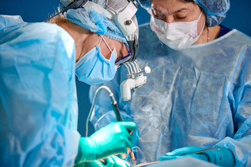 Surgeon performing cosmetic surgery in hospital operating room. Surgeon in mask wearing loupes...