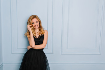 Portrait of joyful young blonde woman wearing elegant black lace dress. Blue wall on background. Her mouth opened, touching her face. She has long blonde wavy hair.