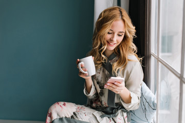 Fototapeta Pretty blonde girl sitting on window sill with cup of coffee, tea and smartphone in hands. She has long blonde wavy hair, smile and looking at her phone. Wearing beautiful silk pajama. obraz