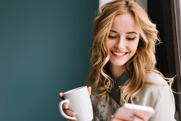 Young blonde woman with wavy hair looking at her phone and holding cup in hand, happy morning messages. Wearing nice pajama. Room with blue, turquoise wall on background. 