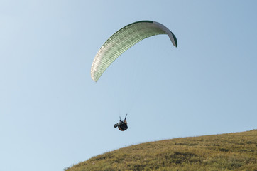A man flies on a paraglider over green fields in the hot summer. Against the background of blue sky...