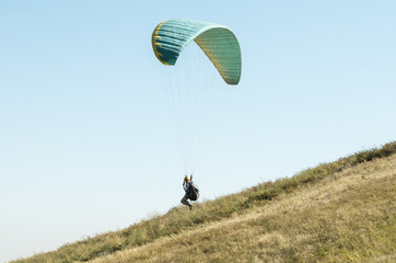 A man flies on a paraglider over green fields in the hot summer. Against the background of blue sky and rare clouds