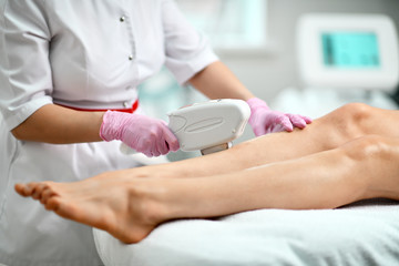 Closeup. Cosmetologist in white gown and gloves performes a procedure on patient leg with medical equipment laser in equiped beauty parlor