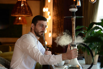 Bearded man in a white shirt with short brown hair smokes a beige wooden hookah with lightning patterns in the lounge bar. The interior of the restaurant with flowers. The concept of rest.