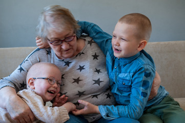 elderly woman sits and plays with her grandchildren on couch. Children love grandmother very much. Boys kiss and hug their beloved grandmother