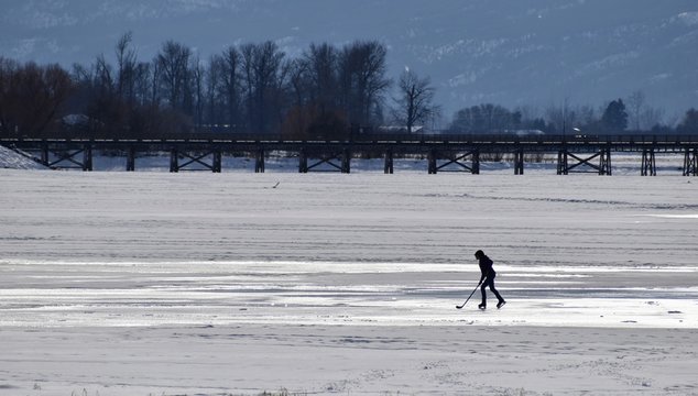 Silhouette of teenage youth alone on a frozen lake playing hockey, ice shining, Wooden pier with trees in the background.. Theme is fitness, practice, solitude. Salmon Arm, British Columbia - Powered by Adobe