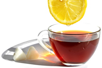 Glass cup of tea with long rainbow shadow in morning rays of sunlight, pieces of sugar in shape of heart and slice of sour eellow lemon with drop of refreshing juice on white background. Isolated