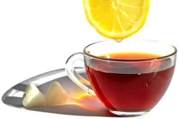 Yellow slice of sour lemon with drop of refreshing juice over glass cup of tea with long rainbow shadow in morning rays of sunlight and pieces of sugar in shape of heart on white background. Isolated