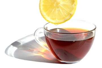 Yellow slice of bitter sour lemon with a drop of refreshing juice over transparent glass cup of hot black tea with long rainbow shadow in morning rays of sunlight on white background. Isolated