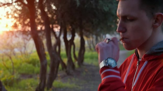 Camera follows the hand with cigarette of handsome young man smoking in the park at sunset. Face of smoking guy in slow motion