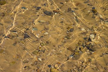 Transparent water surface with ripples through which the sandy bottom with pebbles is visible