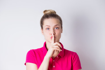 Beautiful young female in pink dress demonstrates silence gesture, keeps forefinger on lips on white background.