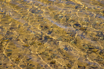 Transparent water surface with ripples through which the sandy bottom with pebbles is visible
