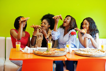 Four young african girls in bright colored restaurant eating pizza slices in hands.