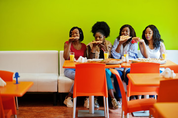 Four young african girls in bright colored restaurant eating pizza slices in hands.