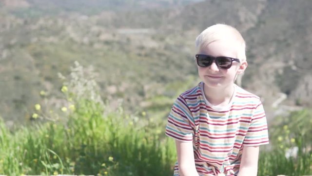 A handsome albino boy in sunglasses sits against a background of mountains. The boy smiles. It's sunny outside.