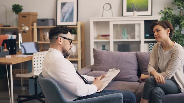 Professional psychologist bearded young man is discussing problems with depressed woman in office. Specialist is asking questions sitting in armchair holding paper.