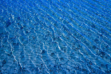 Blue water surface with ripples through which the sandy bottom with pebbles is visible