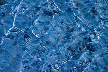 Blue water surface with ripples through which the sandy bottom with pebbles is visible