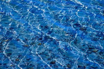 Blue water surface with ripples through which the sandy bottom with pebbles is visible