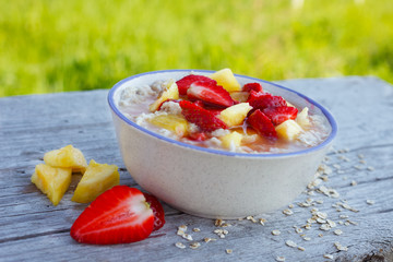 Oatmeal porridge in bowl with fresh strawberry and pineapple. Outdoor image, green meadow in background. Healthy eating.