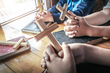 Obraz na płótnie Canvas Christian children group holding christian cross and praying together around wooden table with open bible page at home, prayer meeting concept.