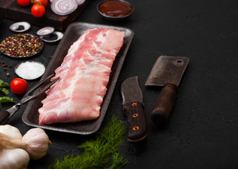 Plastic tray with raw pork ribs and vintage knife and fork on black stone background. Fresh tomatoes and red onion with garlic salt and pepper.