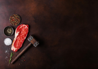 Raw sirloin beef steak with meat hammer on rusty background. Salt and pepper with fresh rosemary and bowl of oil.Space for text