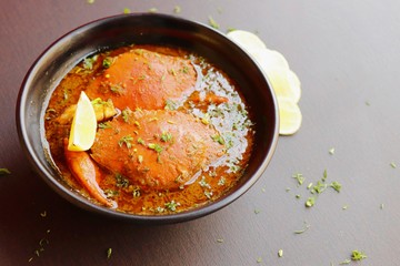 Indian Cuisine, Spicy Crabs Curry in red gravy, garnished with fresh coriander and lemon wedges or lemon slices on dark moody background