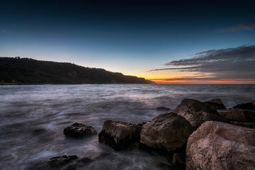 tramonto sul mare alla baia Flaminia di Pesaro con scogli in primo piano e collina sullo sfondo
