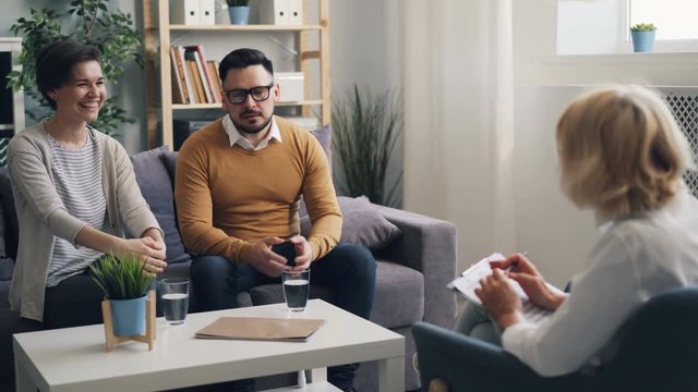 Joyful couple man and woman are smiling talking to psychotherapist during family counseling in modern clinic. Happy clients and successful doctor concept.