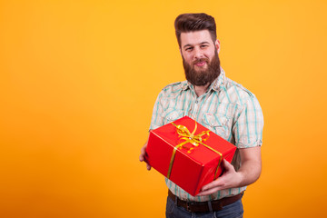 Young bearded man looking at the camera and holding a gift box over yellow background