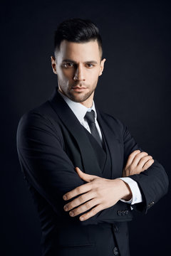 Handsome confident man in black suit with arms crossed on dark background