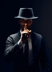 Serious man in black suit and hat keeps finger on lips, making hush gesture and keep conspiracy...