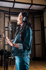 Naklejka premium attractive emotional woman singing near microphone in recording studio